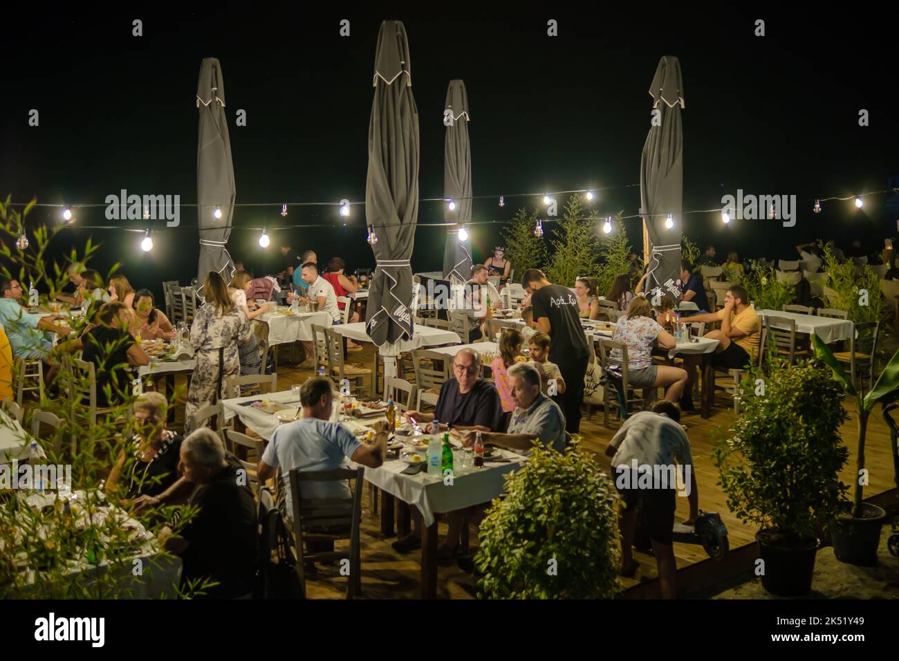 Nea Kallikrateia, Greece - August 28, 2022 :  Greeks and tourists enjoying dinner outdoors at a cozy illuminated restaurant in front of the sea Stock Photo