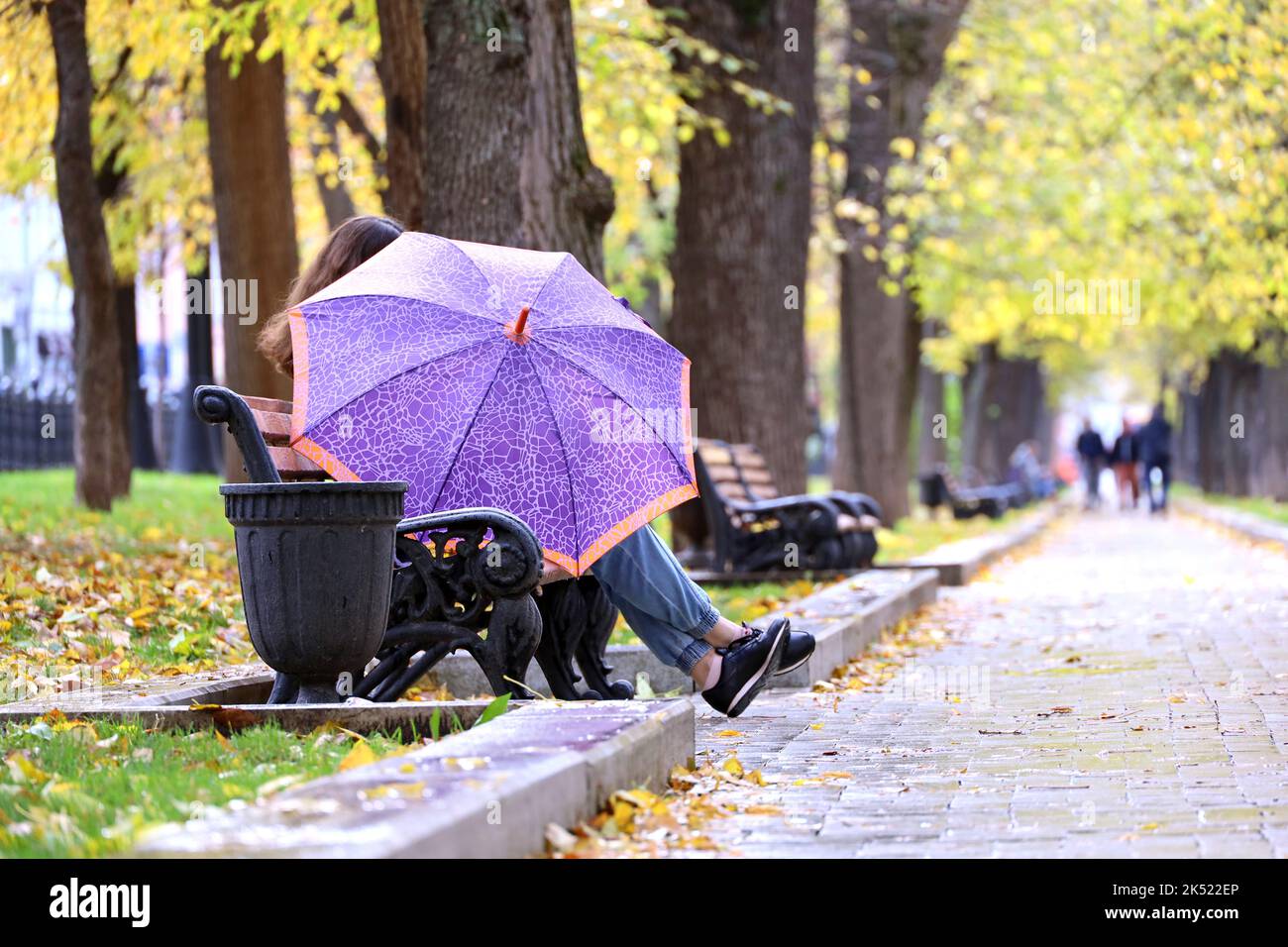 Rain in autumn season, girl with umbrella sitting on a bench in city park Stock Photo