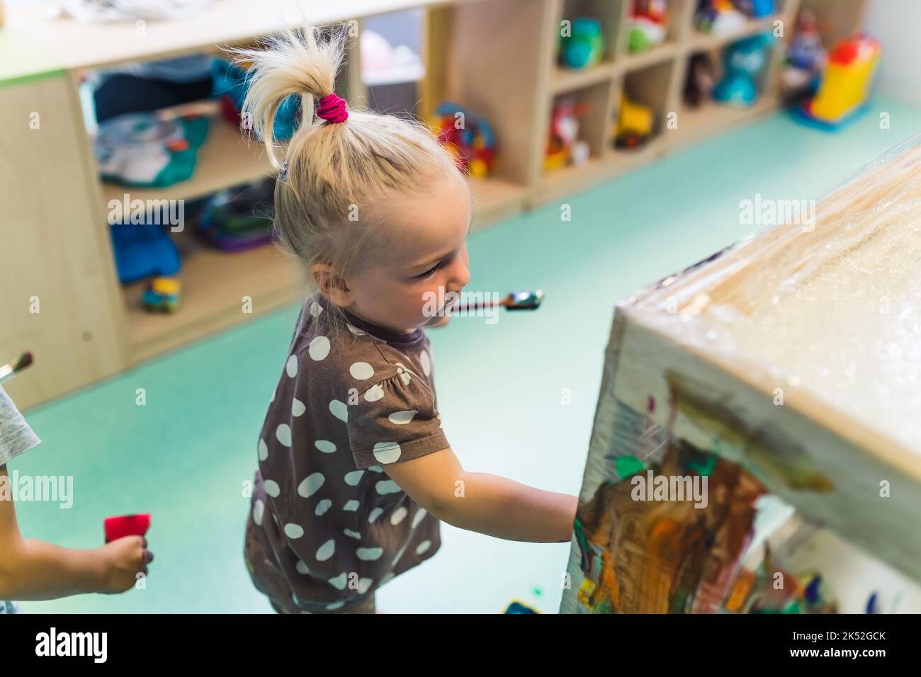 sweet little girl with blond hair having fun and painting with her friends in the nursery, medium shot shelves full of toys in the background. High quality photo Stock Photo