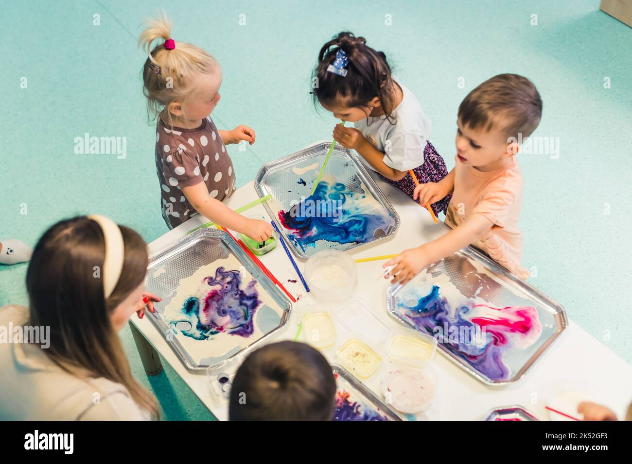 kids standing at the table full of watercolors and trying to paint with their teacher, nursery kids' concept. High quality photo Stock Photo