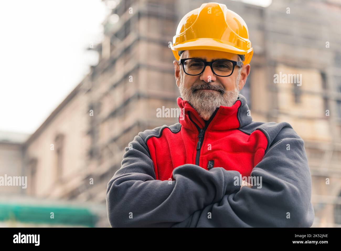 Proud caucasian male blue-collar worker looking straight to the camera with arms crossed in front of his chest. Yellow protective helmet. Construction site in the background. High quality photo Stock Photo