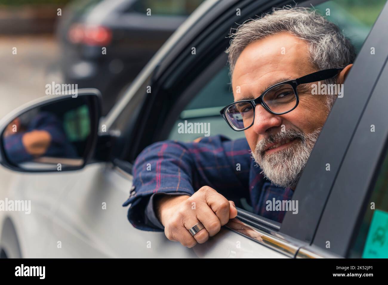 Friendly caucasian man with gray hear, gray beard, and black glasses looking out of the driver's window and looking at camera with a smile. Closeup outdoor shot. Commuting with a car. High quality photo Stock Photo