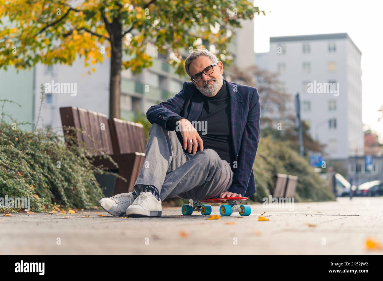 Proud and confident caucasian gray-haired man in his 60s dressed in business casual way sitting cross-legged on modern neon orange skateboard and looking at camera. Sport for everyone. High quality photo Stock Photo