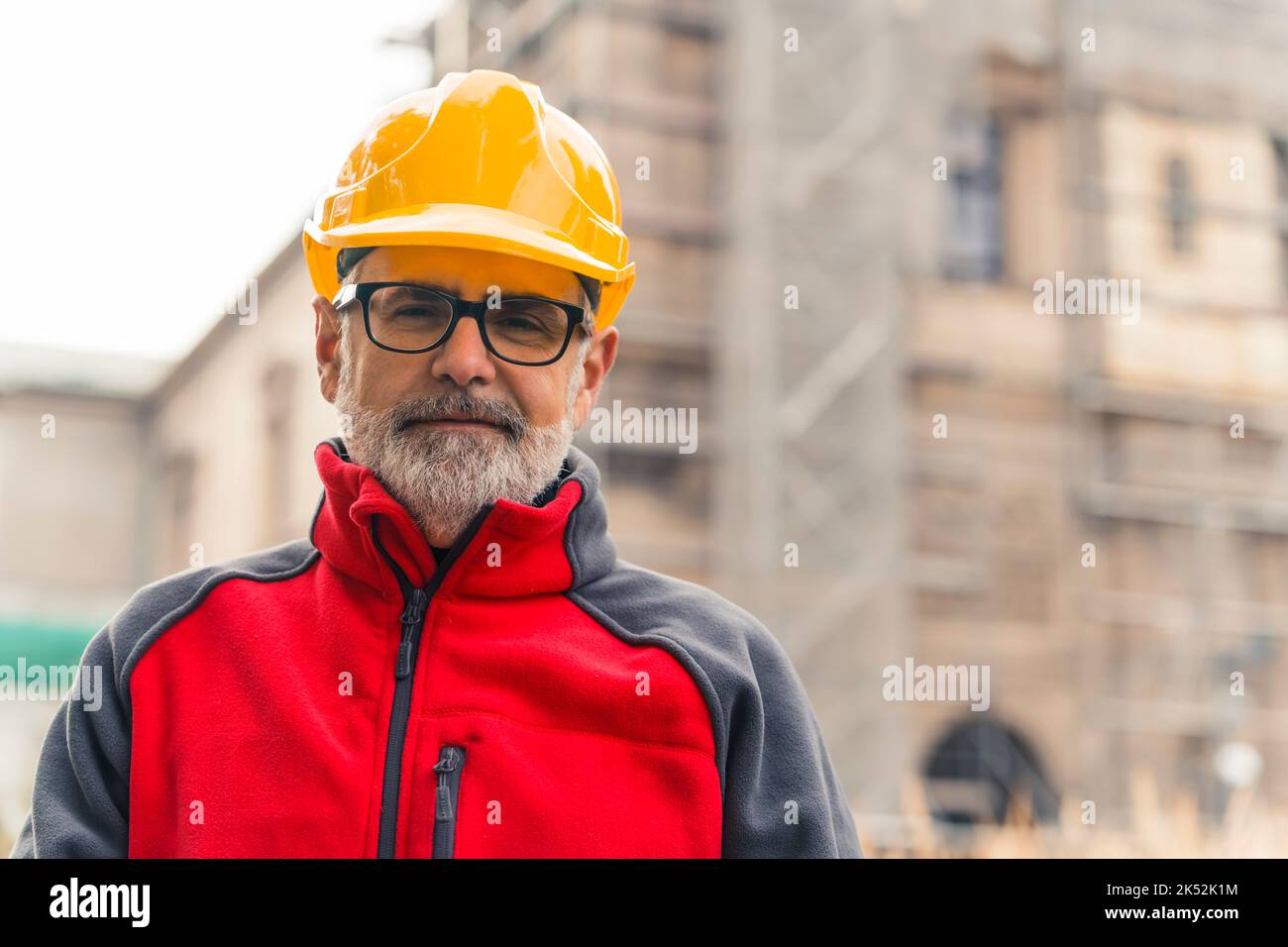 Satisfied with his work caucasian construction worker in his late 50s looking at camera in protective gear and standing in front of blurred building during renovation. Blue-collar jobs concept. High quality photo Stock Photo