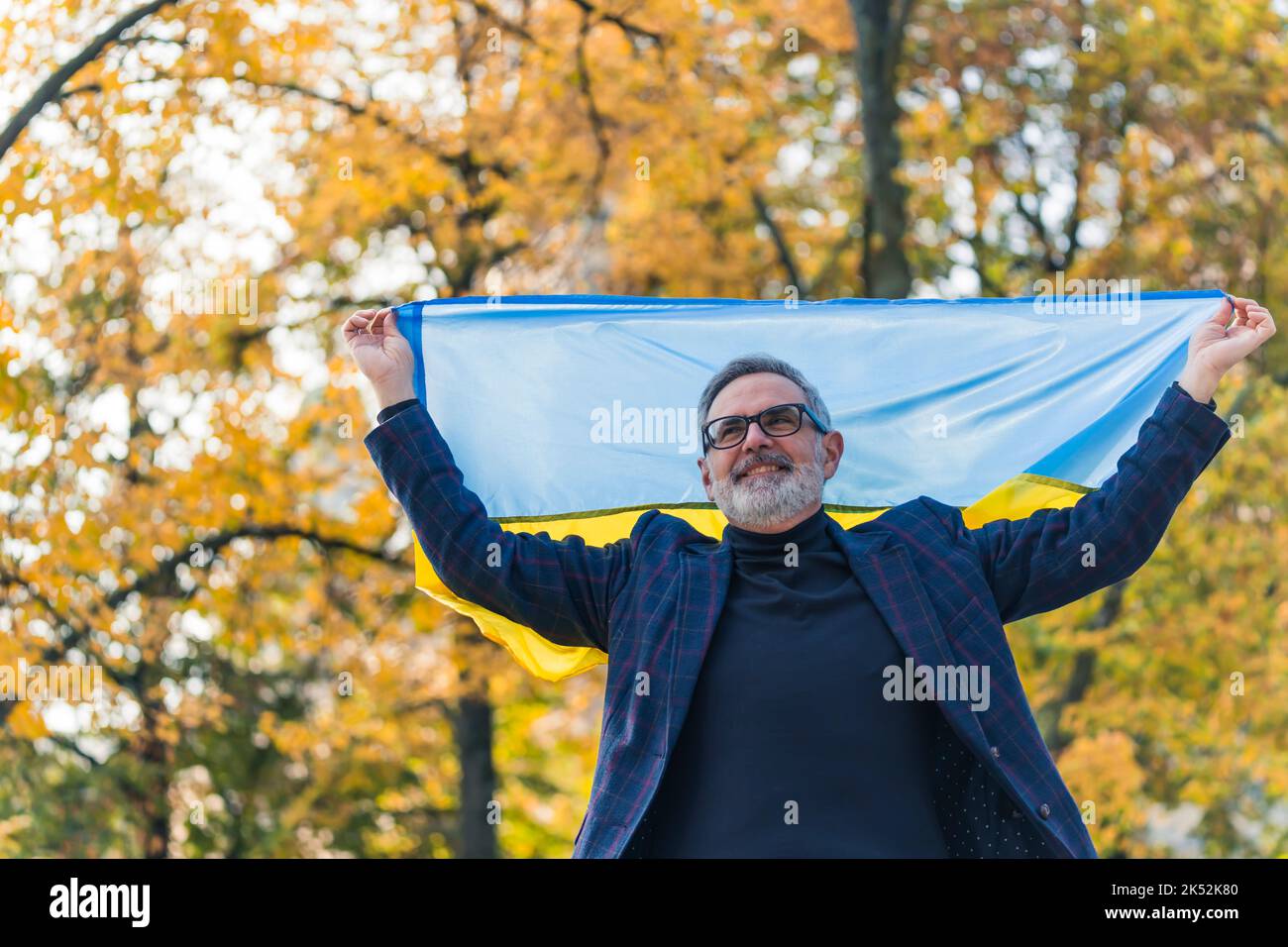 Support for Ukraine. Middle-aged caucasian bearded male European man dressed in business casual way, walking outside and holding blue-and-yellow Ukrainian flag up in the sky with two hands. Blurred autumn trees in the background. High quality photo Stock Photo