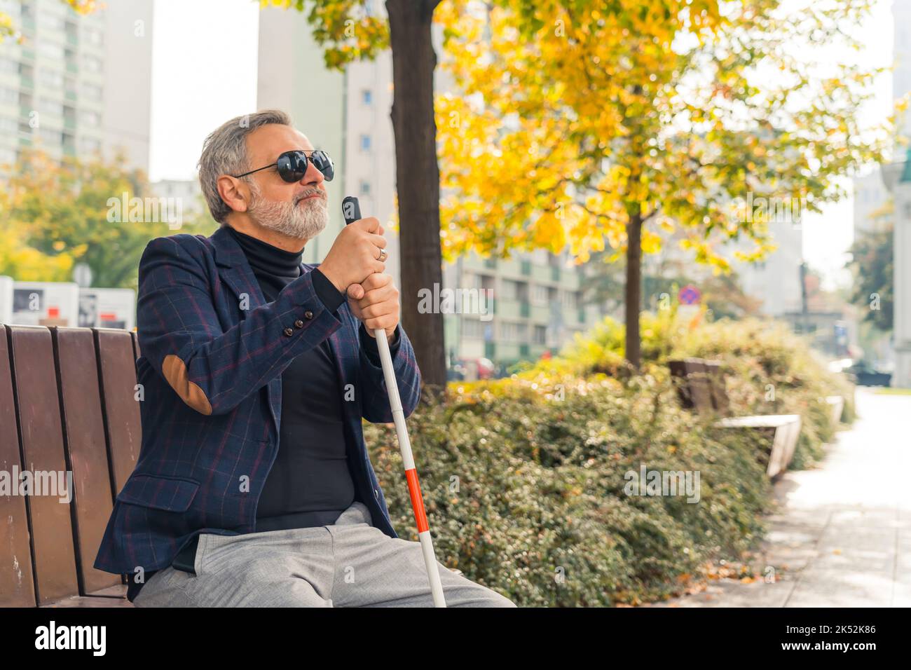 Relax in fresh air. Break from loud city at park. Autumn season. Good-looking caucasian bearded adult man with sight disability sitting on bench, holding his white walking cane, and relaxing. High quality photo Stock Photo