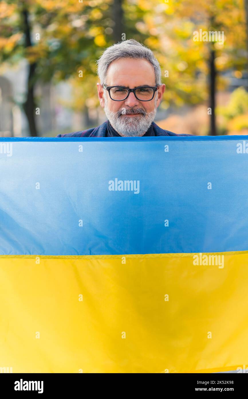 Vertical outdoor shot of adult caucasian gray-haired man in glasses covering his body with blue-and-yellow Ukrainian flag to show support for Ukraine during war. High quality photo Stock Photo