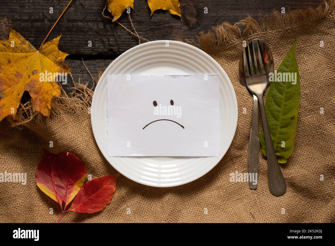 sad smiley is drawn on paper lies on a white plate on a wooden table in autumn yellow leaves, sadness sadness depression, crisis, nothing to eat, bad Stock Photo