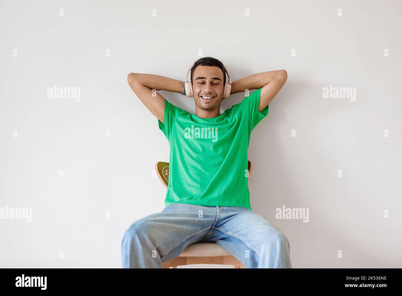 Relaxed arab man listening to music in wireless headphones, resting on chair with hands behind head over white wall Stock Photo