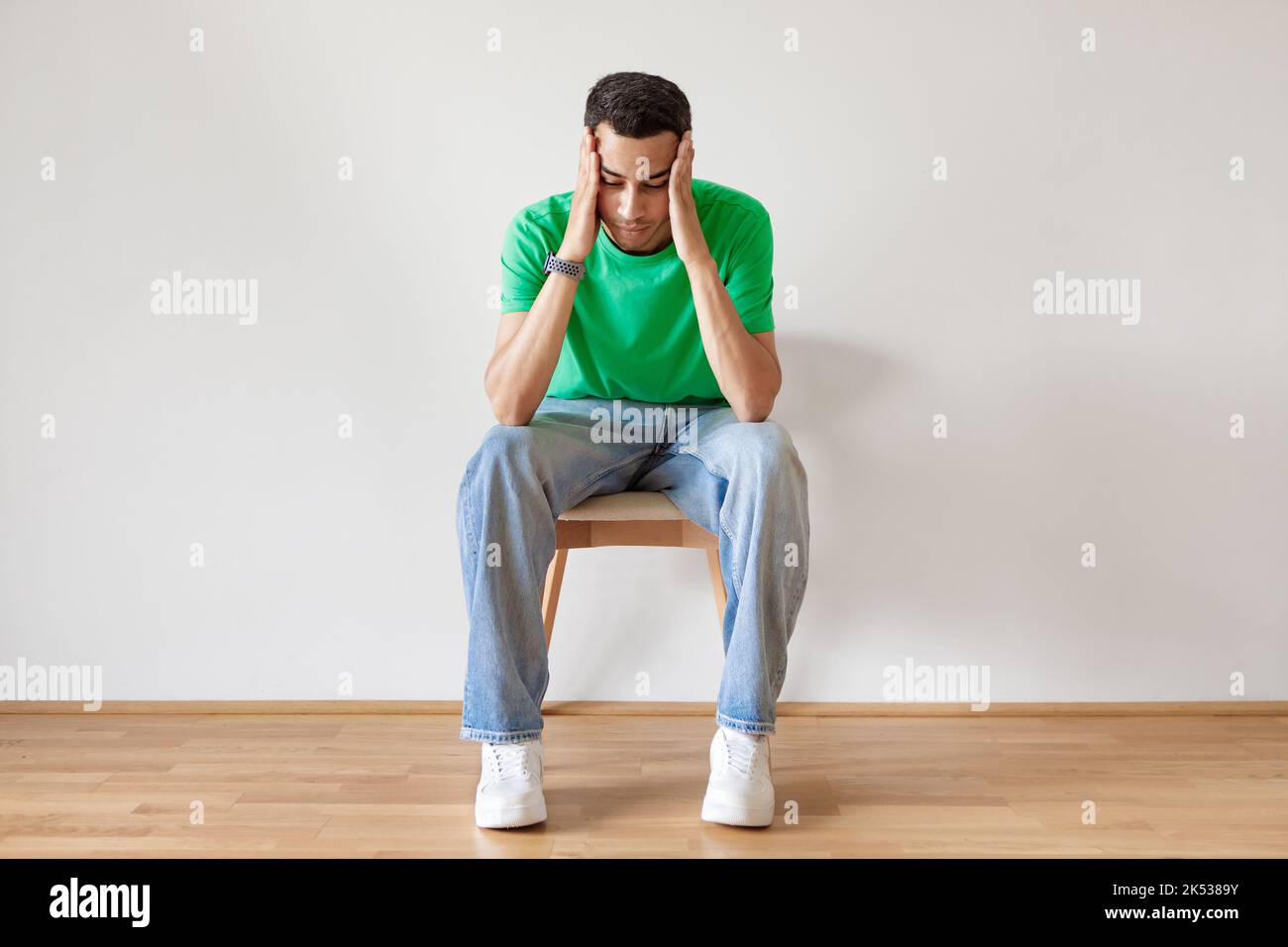 Pensive young arab man thinking, touching head with both hands, sitting on chair over light wall, copy space Stock Photo