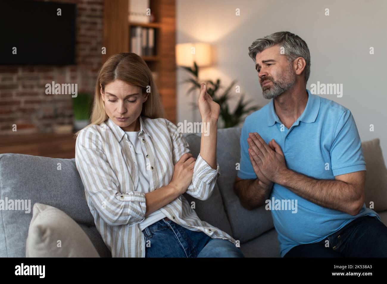 Sad mature caucasian husband begs wife for forgiveness, lady ignores arguments on sofa in living room interior Stock Photo