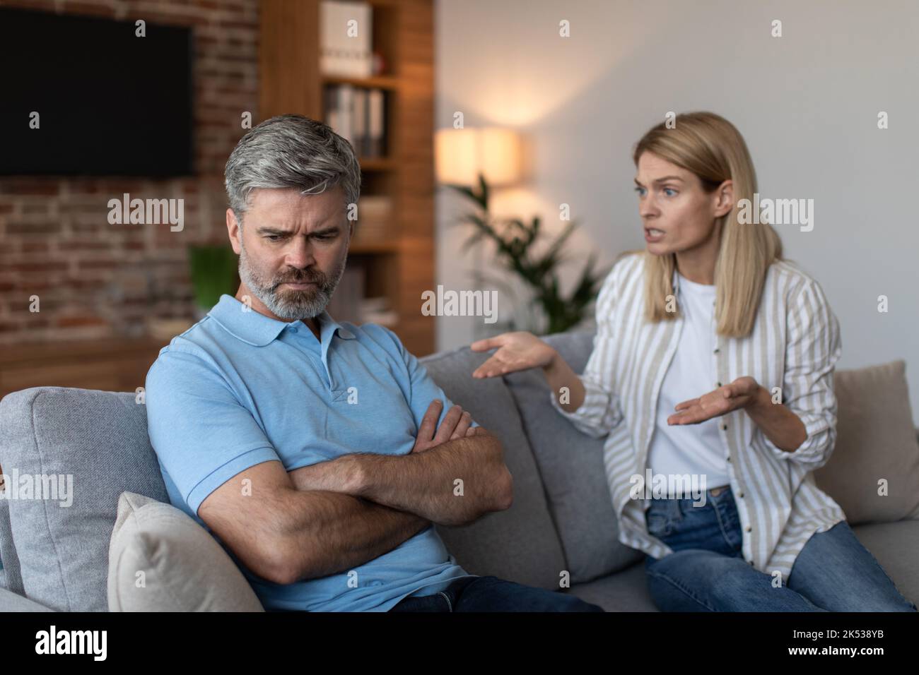 Unhappy mature caucasian wife scolds at offended husband, quarreling on sofa in living room interior Stock Photo