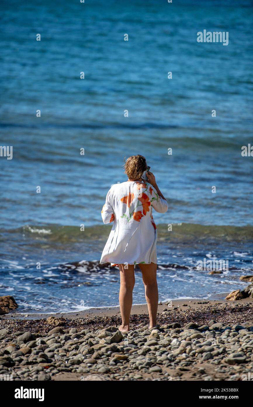 older woman walking along sandy beach using smartphone to make a call while on holiday on the greek island of zante or zakynthos. Stock Photo