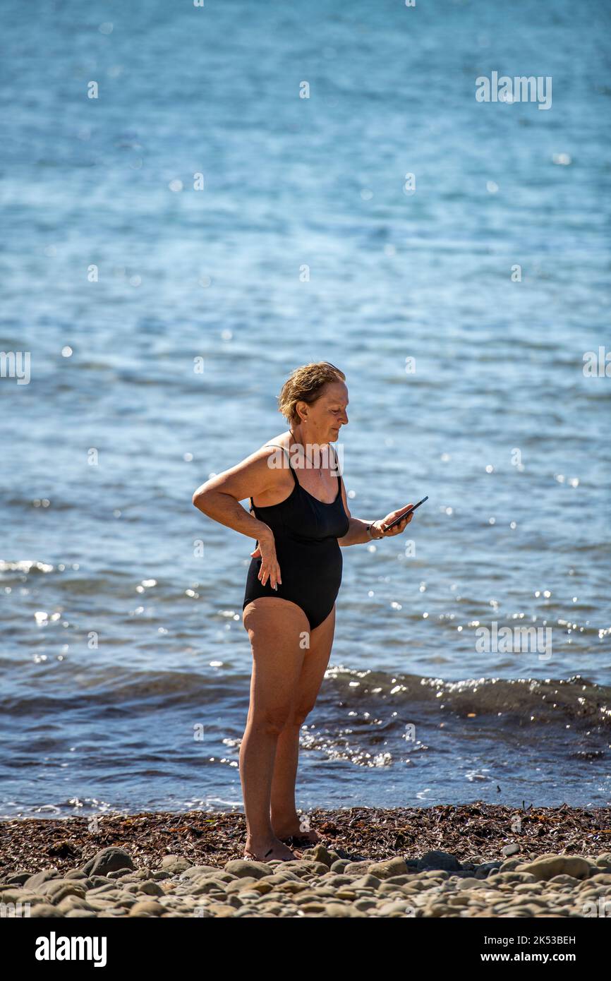 older woman standing on a sunny beach using a smartphone wearing a black swimming costume looking at the phone screen reading Stock Photo