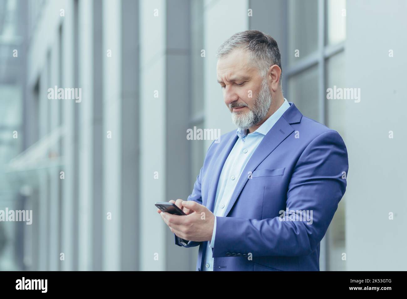 Close-up photo. Senior gray-haired man, a businessman in a suit, stands near the office center and calls a taxi from the phone, typing the number, waits, is in a hurry to meet. Stock Photo