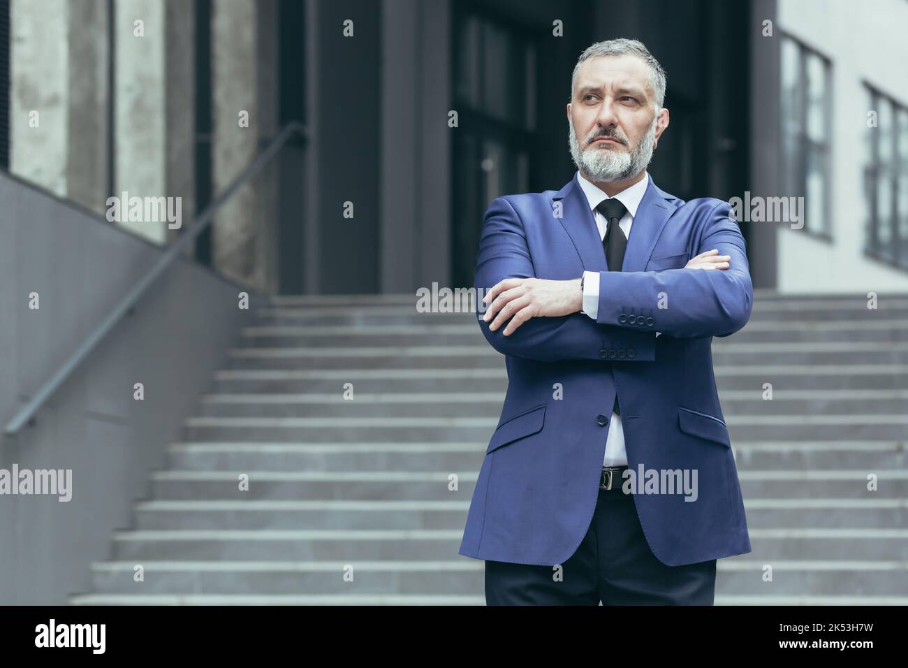 Portrait of a senior gray-haired man director, ceo on the background of the stairs of the office center. He is standing serious in a suit, arms crossed in front, looking to the side. Stock Photo