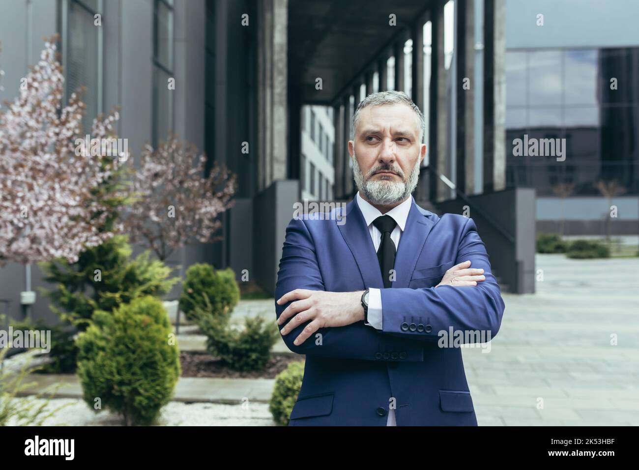 Portrait of a senior gray-haired man director, ceo on the background of the office center and trees. He is standing serious in a suit with tie, arms crossed in front, looking to the side. Stock Photo