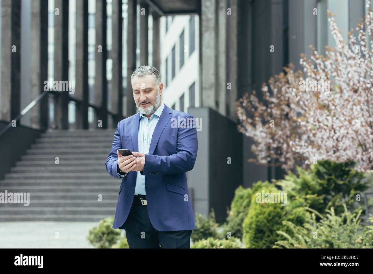 Senior gray-haired man, a businessman in a suit, stands near the modern office center and calls a taxi from the phone, typing the number, waits, is in a hurry to meet. Stock Photo