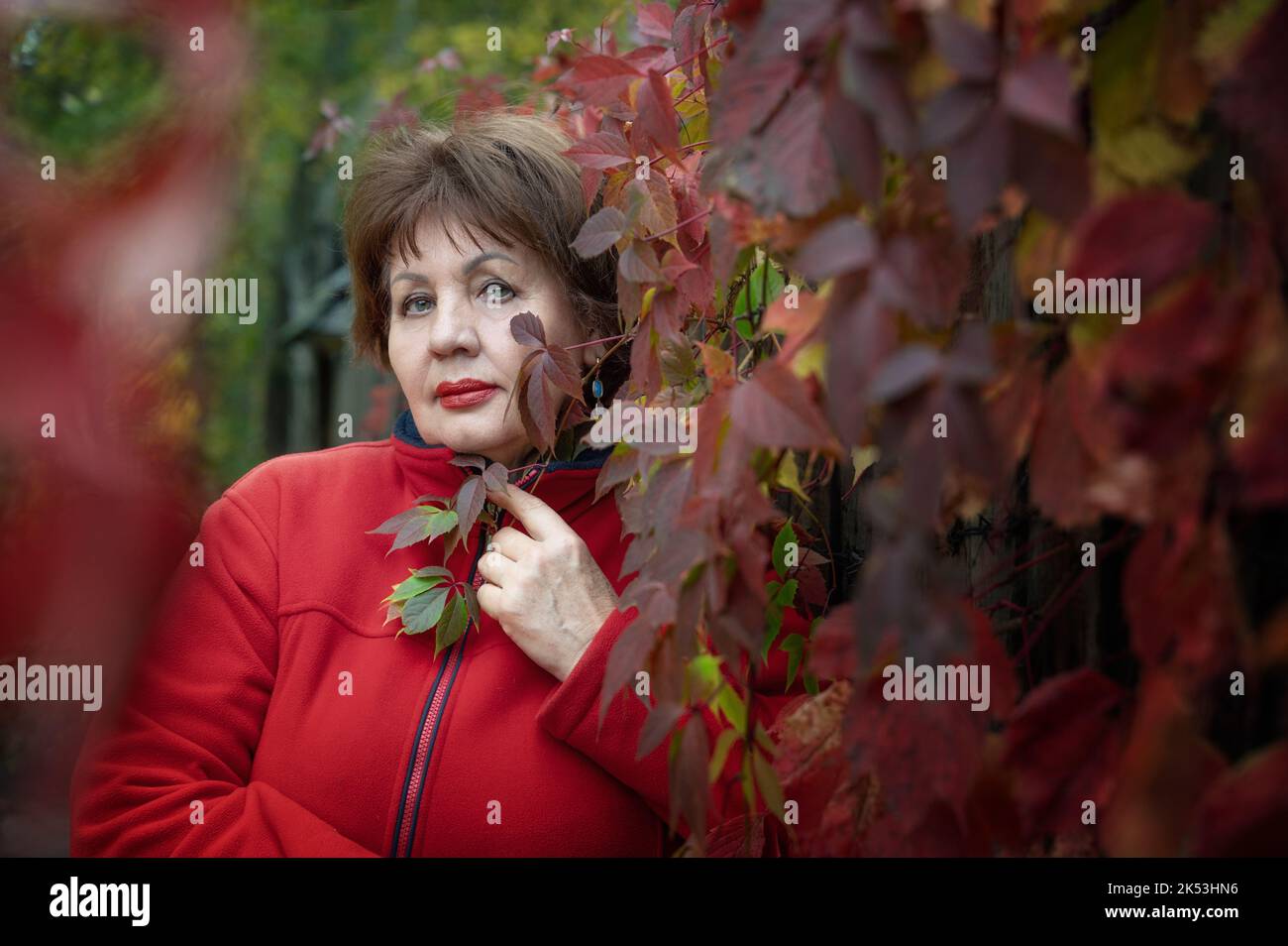A beautiful brown-haired woman of 70 years old is sad in the autumn garden. Portrait of a woman against a background of beautiful autumn bright greene Stock Photo