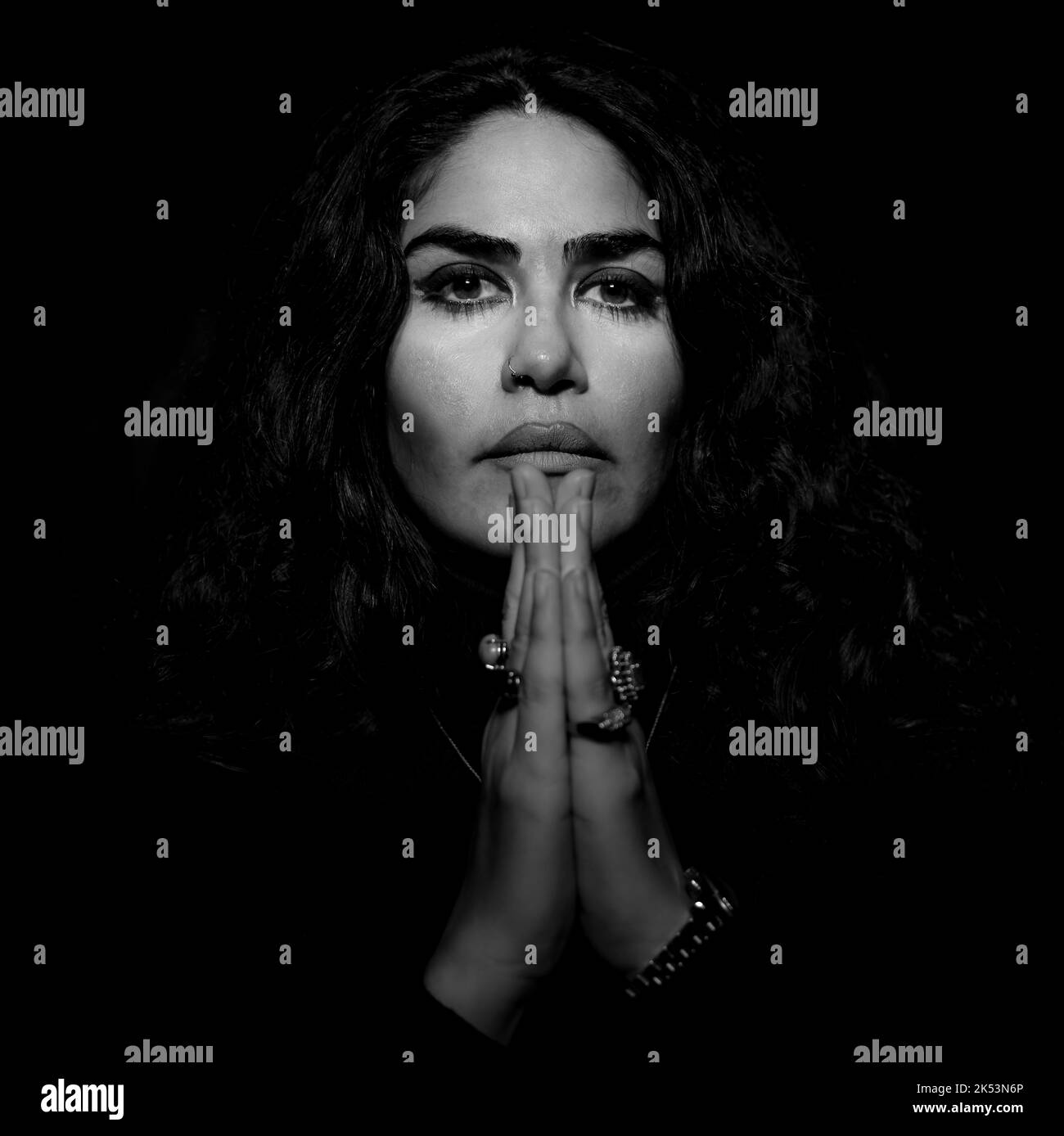 Frontal close-up black and white portrait of brunette Persian woman with long hair in black dress, folded her hands in front of her face, prayer. Stock Photo