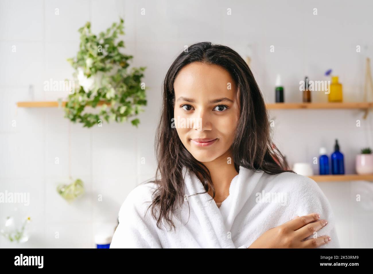 Portrait of young beautiful woman with dark skin in white bathrobe with cosmetic bottle on the background. Confident young woman feeling harmonous and calm in her body. Wellness and body positivity Stock Photo