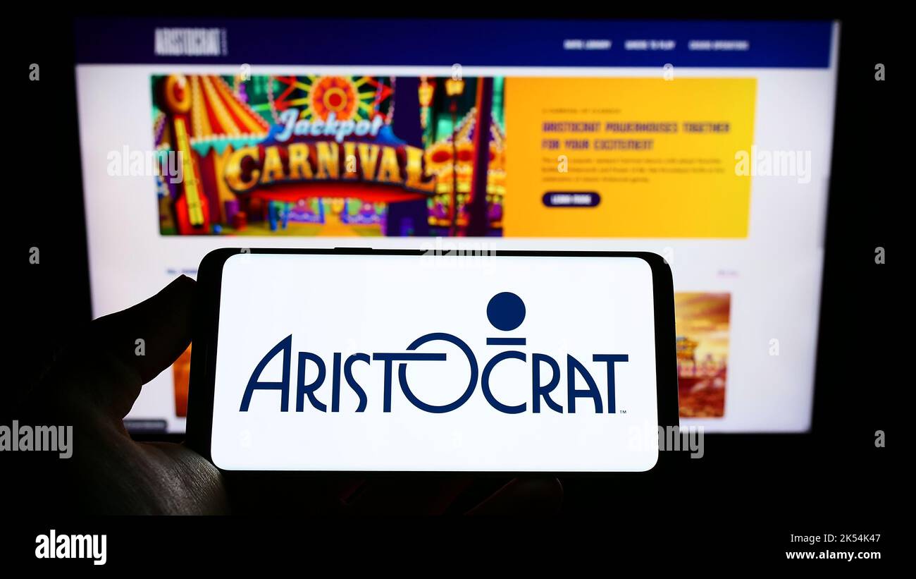 Person holding cellphone with logo of gambling company Aristocrat Leisure Limited on screen in front of business webpage. Focus on phone display. Stock Photo