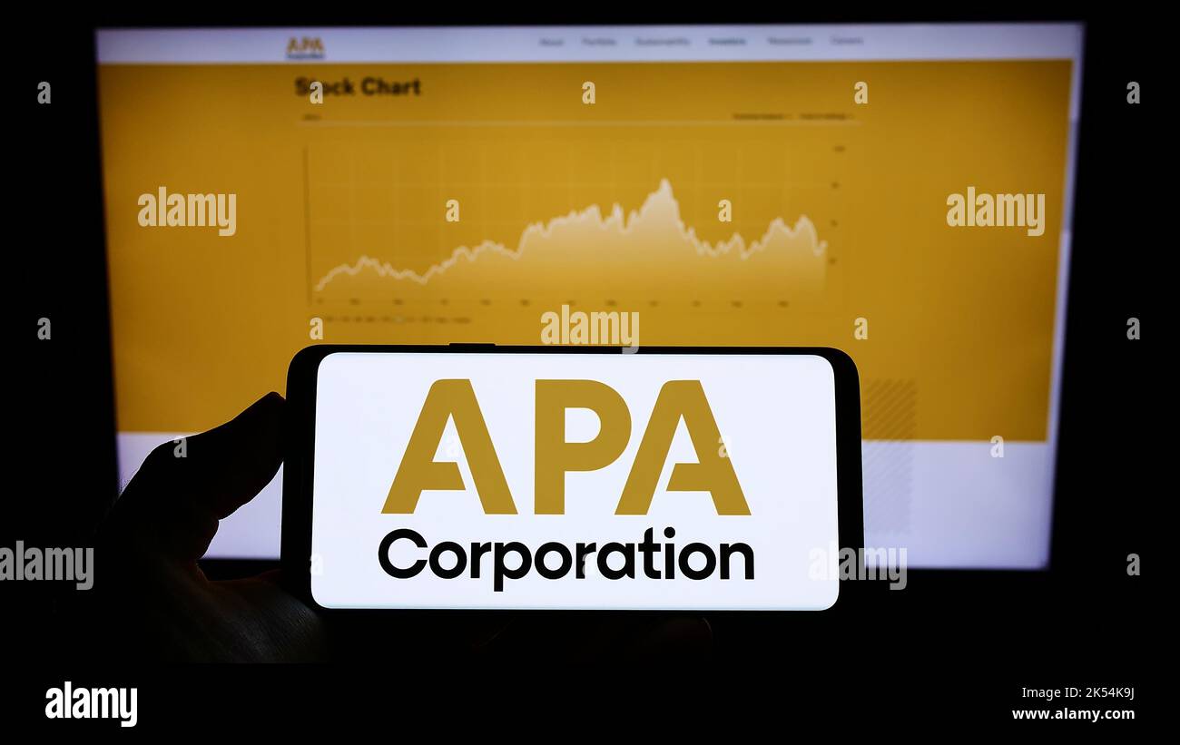 Person holding mobile phone with logo of American petroleum company APA Corporation on screen in front of web page. Focus on phone display. Stock Photo