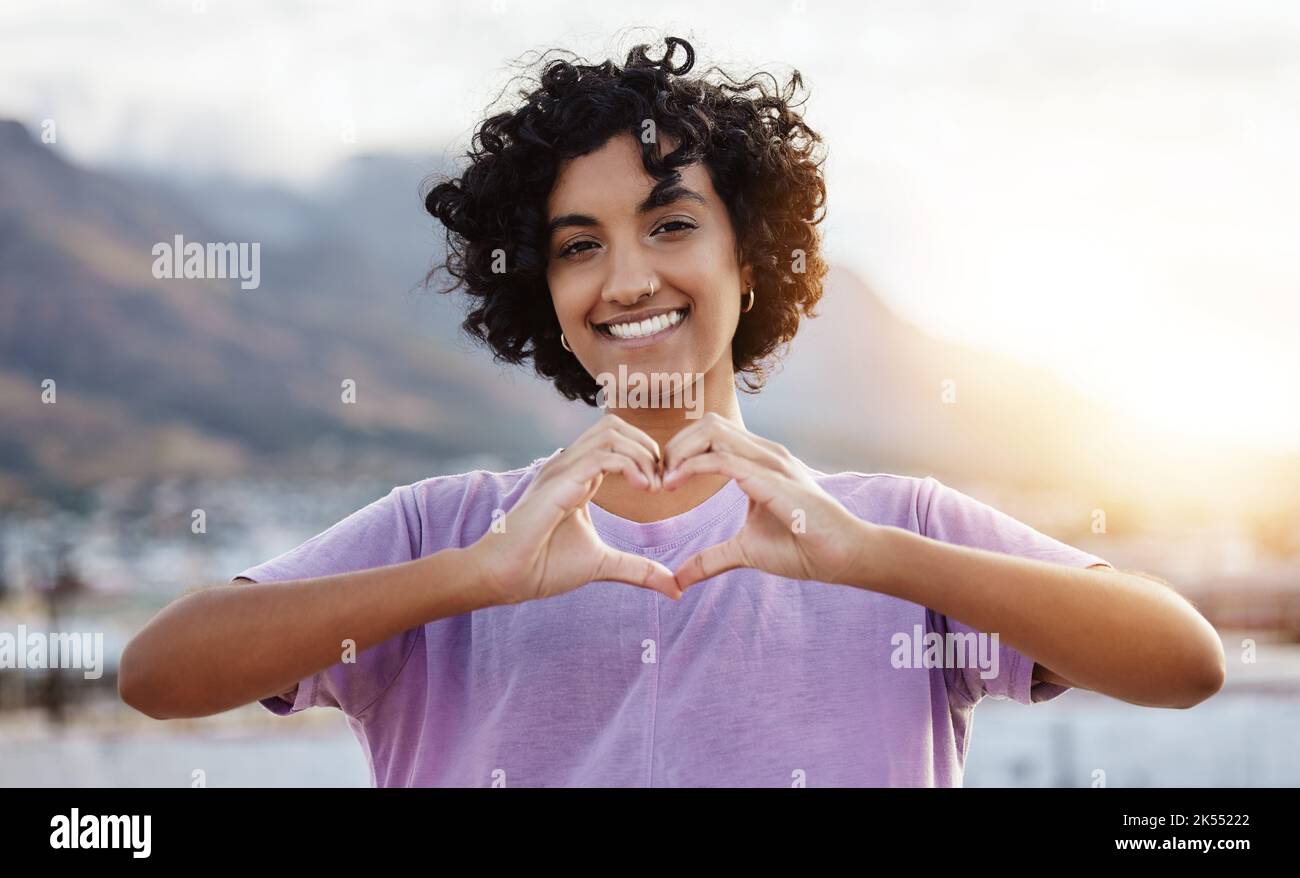 Hand, heart and woman showing love sign in a city, travel, freedom and happy student. Portrait, smile and finger gesture with cheerful indian tourist Stock Photo