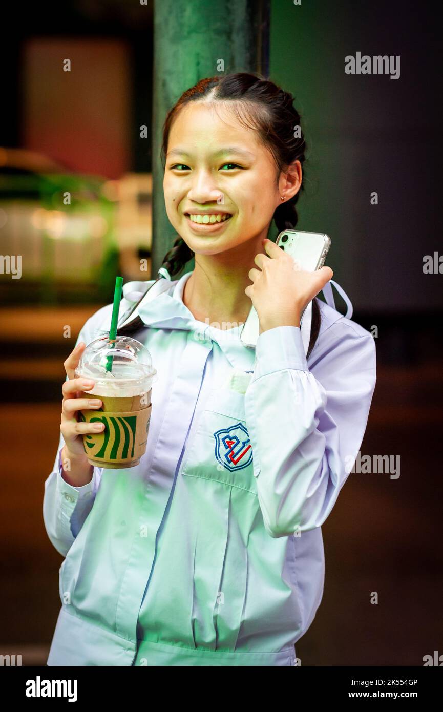 A beautiful Thai school girl poses and laughs holding her Starbucks drink waiting for a bus at Sala Daeng, Bangkok, Thailand. Stock Photo