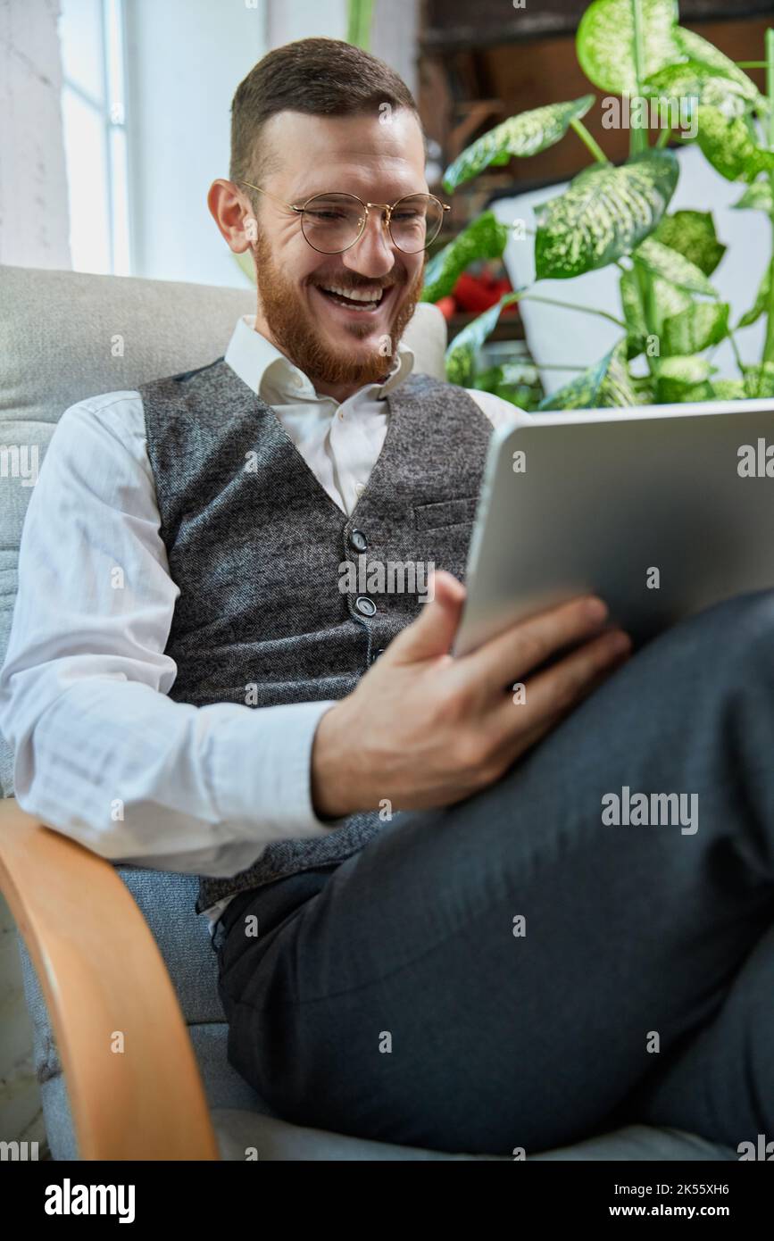 Young relaxed peaceful indian eastern man sitting on coach in modern living room holding tablet computer checking social media, surfing internet Stock Photo