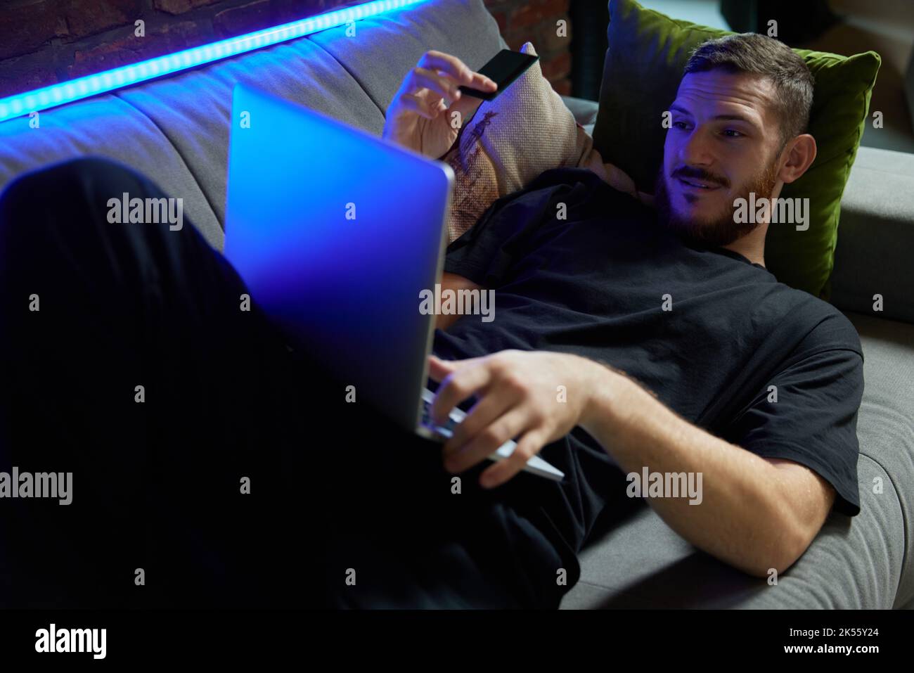 Cheerful handsome caucasian man lying on the sofa with laptop and credit card in hand in living room at home. Neon light. Online shopping and Stock Photo