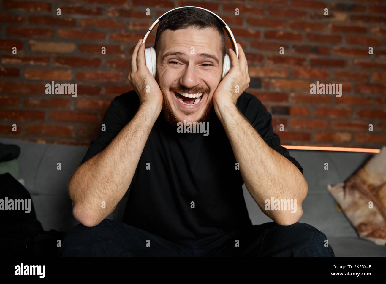 Handsome bearded man in headphones listening to music at home. Music. Half-length portrait of young smiling guy in casual youth style clothes sitting Stock Photo