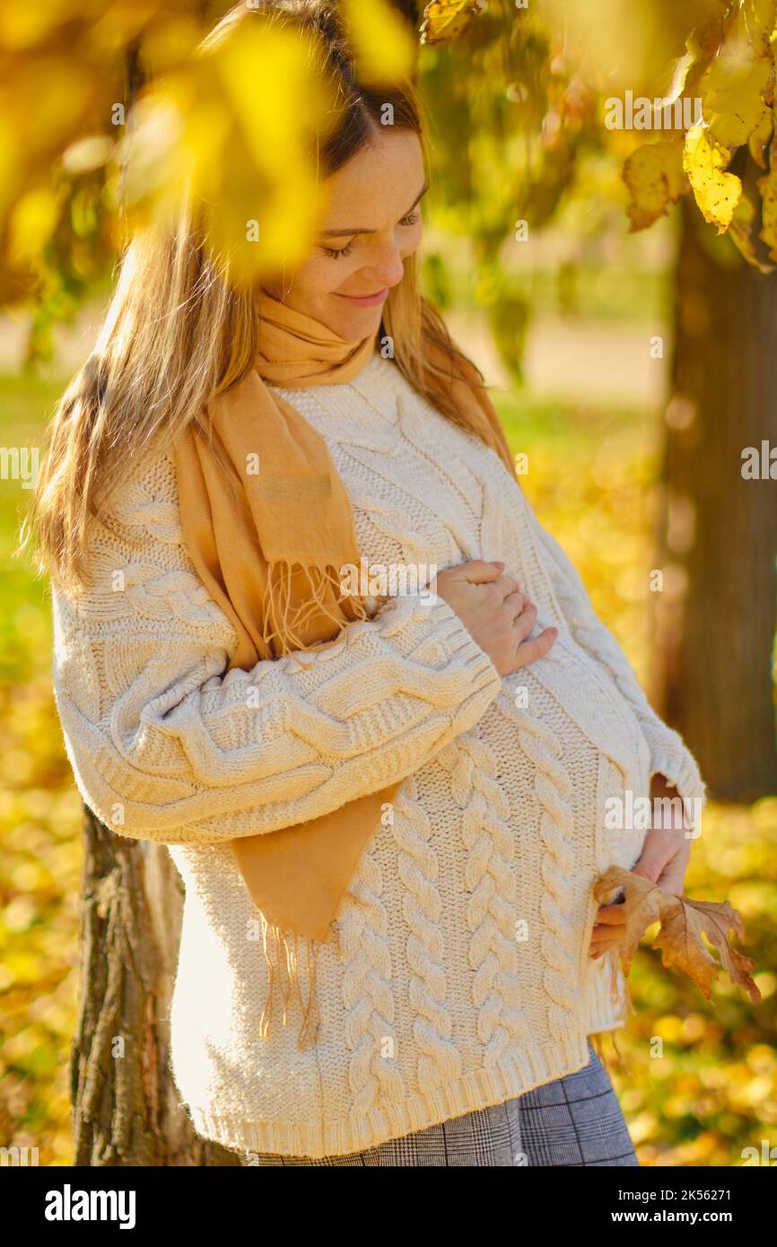 Attractive young pregnant woman with leaf holding her tummy in autumn park. Stock Photo