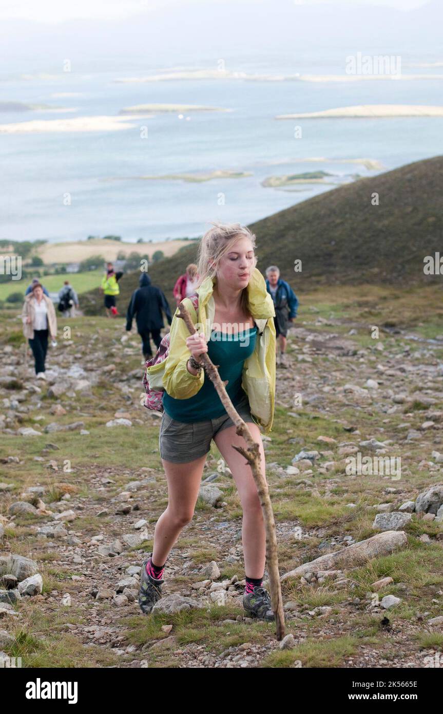 A young lady uses a stick to help her climb Croagh Patrick on Reek Sunday, when Roman Catholic pilgrims climb to the top to follow the steps of Saint Patrick. Stock Photo