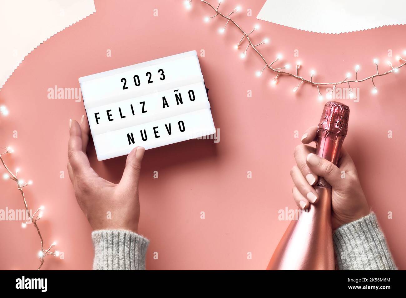 Text Happy New Year 2023 in Spanish Language on lightbox. Light garland, woman hands with lightbox and metallic pink champagne bottle. New Year Stock Photo