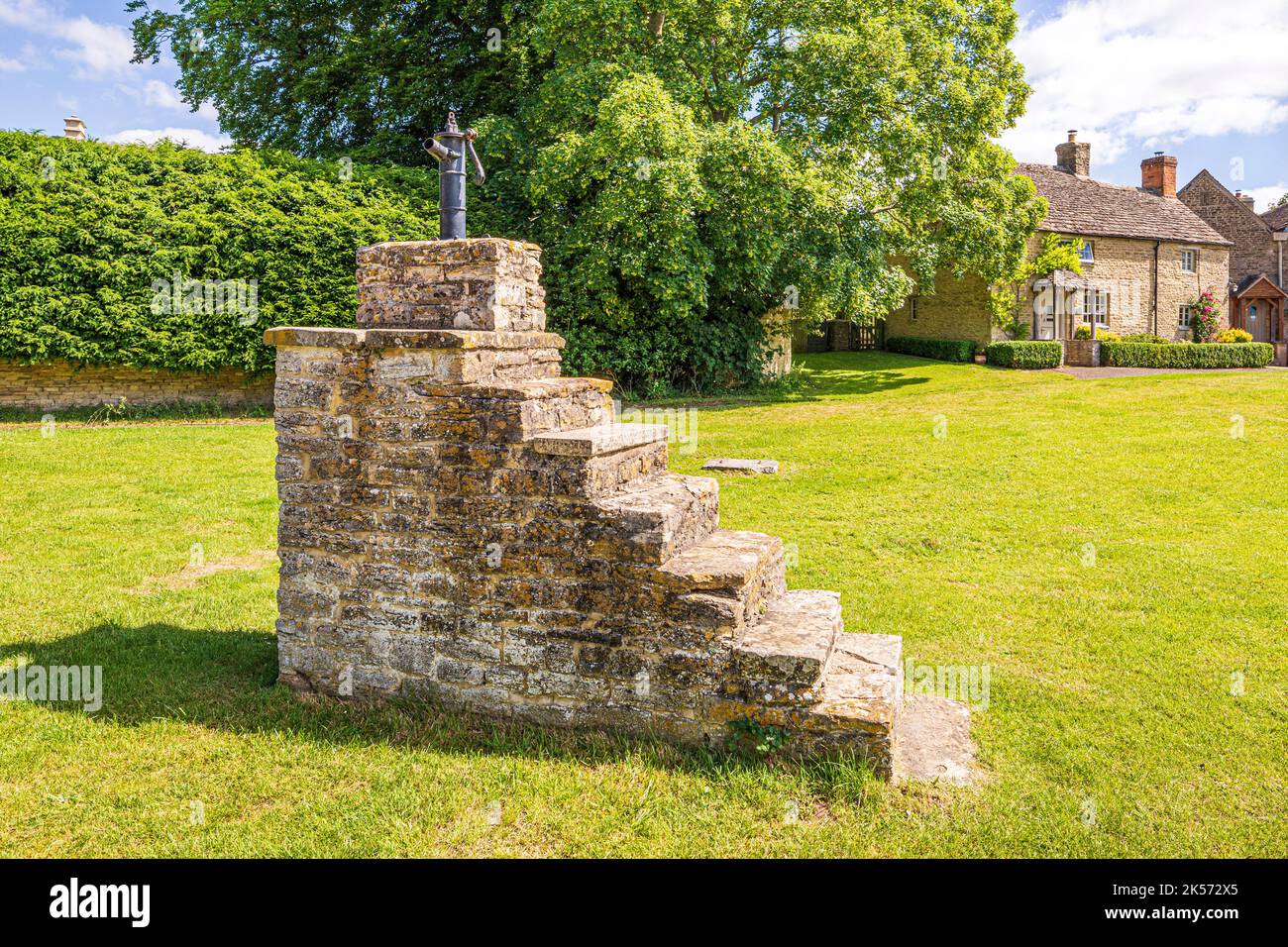 A cast iron water pump on top of a tall stone mounting block on the green in the Cotswold village of Meysey Hampton, Gloucestershire, England UK Stock Photo