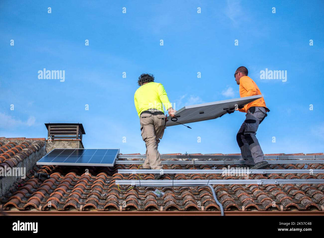 Roof with two people installing solar panels and a blue sky with white clouds in the background. Stock Photo