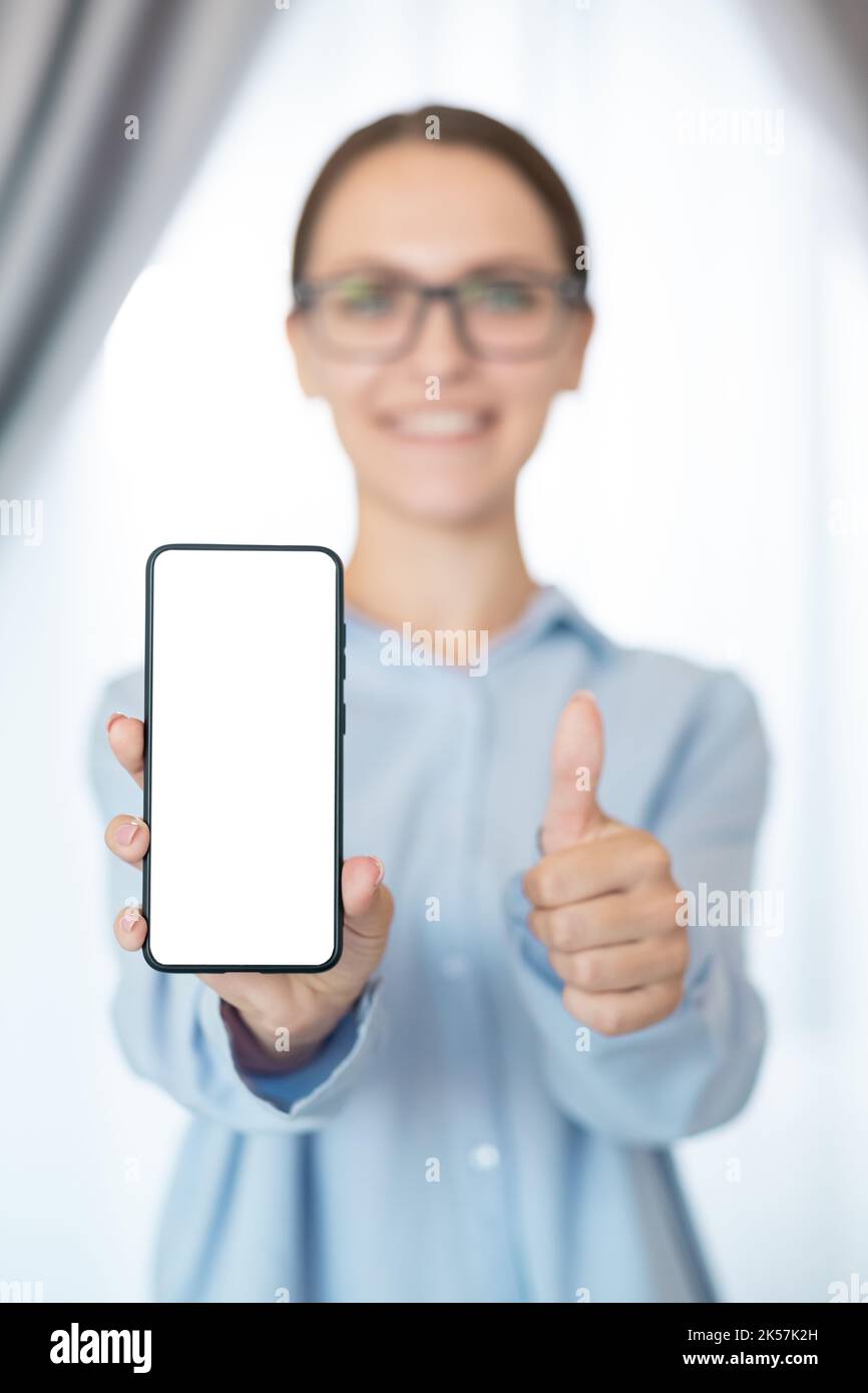 App advertising. Social media marketing. Mobile business. Satisfied cheerful woman recommending white blank mockup phone screen with thumb up like ges Stock Photo