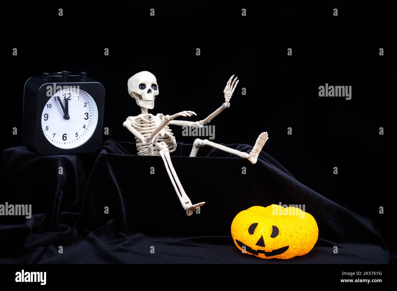 Halloween time. A skeleton crawling out of the grave and waving his hand, greeting, pumpkins with grimaces, a clock on a black background. The eve of Stock Photo