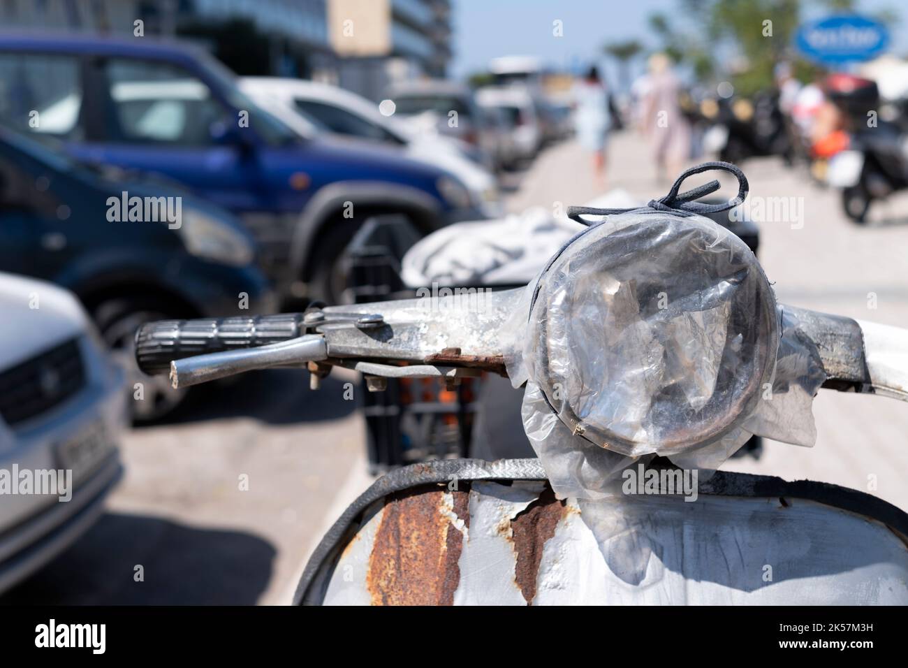 An old rusty motor scooter is parked on the roadside in Greece. The scooter has a broken headlight lens that is temporarily repaired with plastic bag Stock Photo