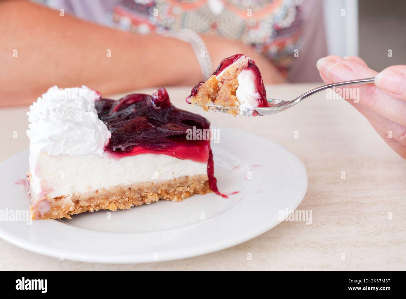 A woman eating a cherry cheesecake. She has a portion of the cheesecake on a fork in front of her Stock Photo