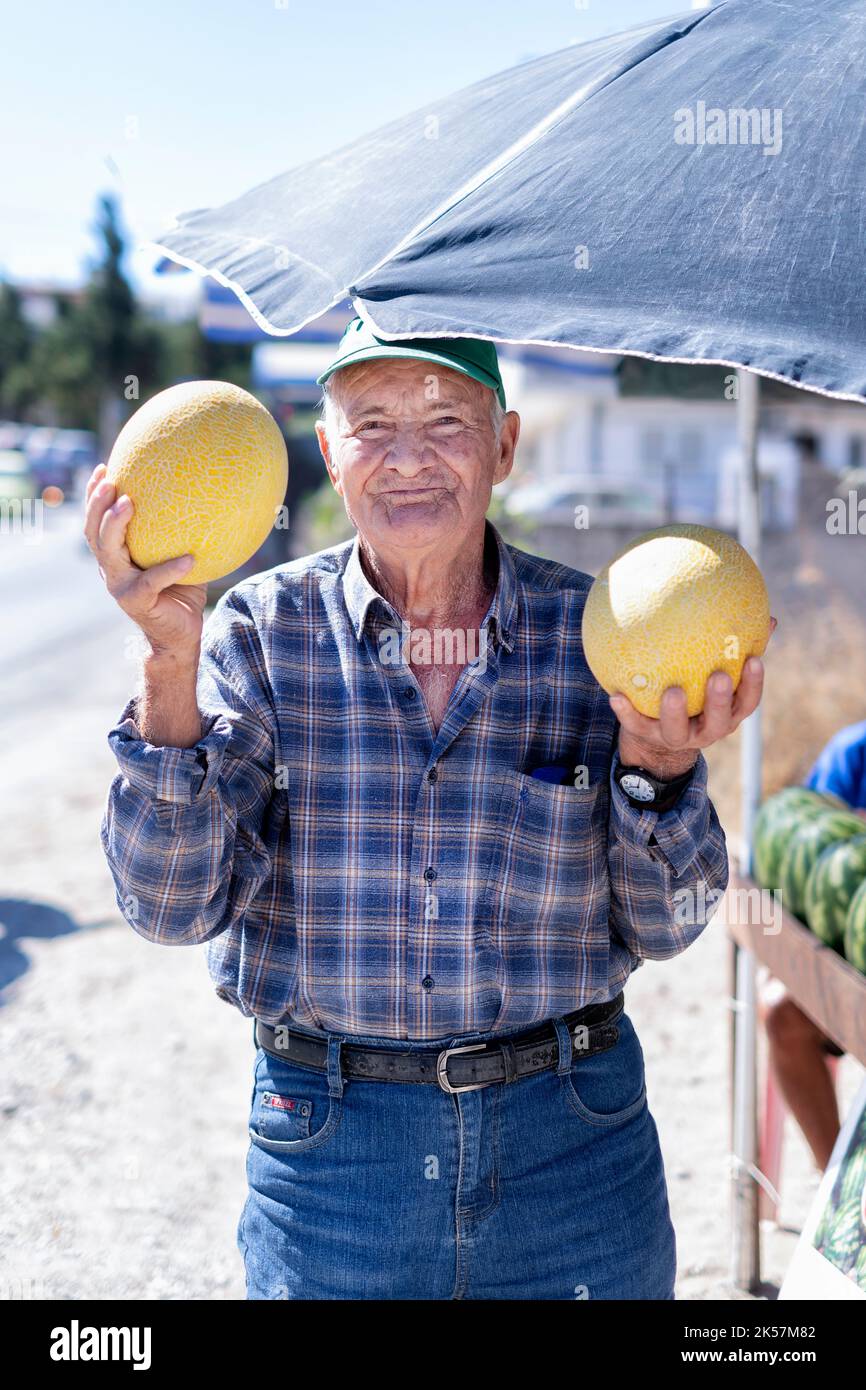An elderly man holding two large yellow honeydew melons Cucumis melo. The man is selling the melons from a stall at the side of the road in Rhodes Stock Photo