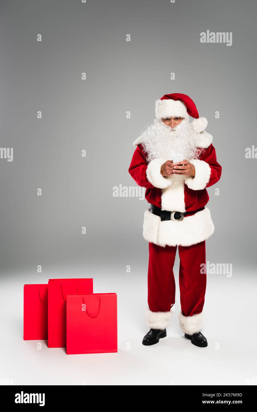 Full length of father christmas using cellphone near shopping bags on grey background,stock image Stock Photo