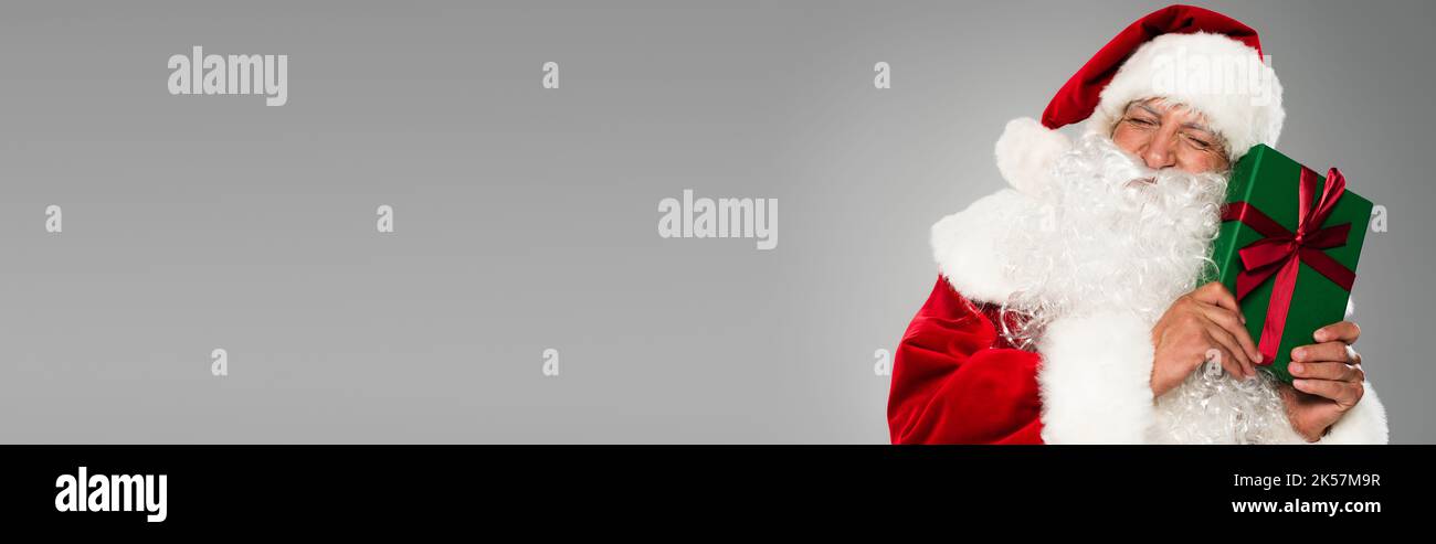 Father christmas in hat closing eyes while holding gift with bow isolated on grey, banner,stock image Stock Photo