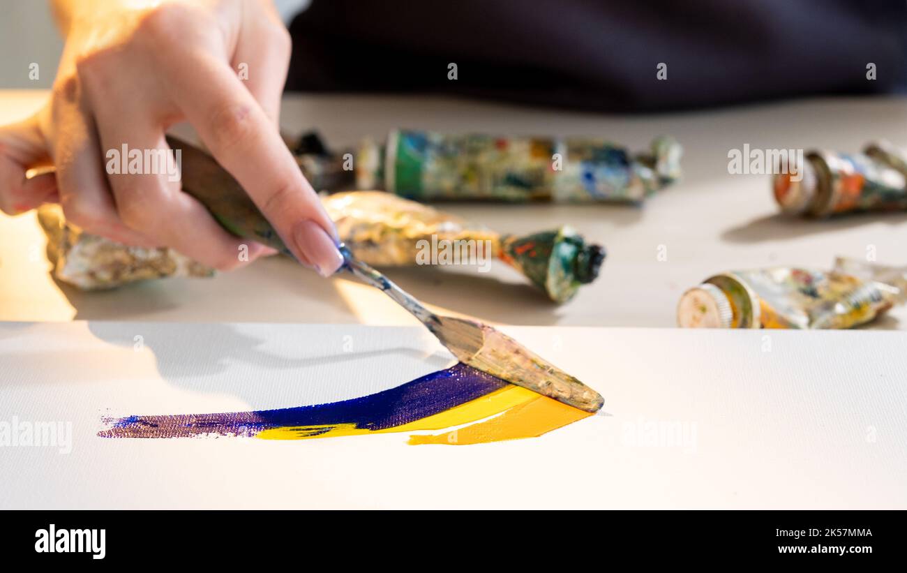Painting lesson. Creative process. Artistic tools. Hobby leisure. Unrecognizable woman mixing blue yellow paint colors with palette knife on canvas. Stock Photo