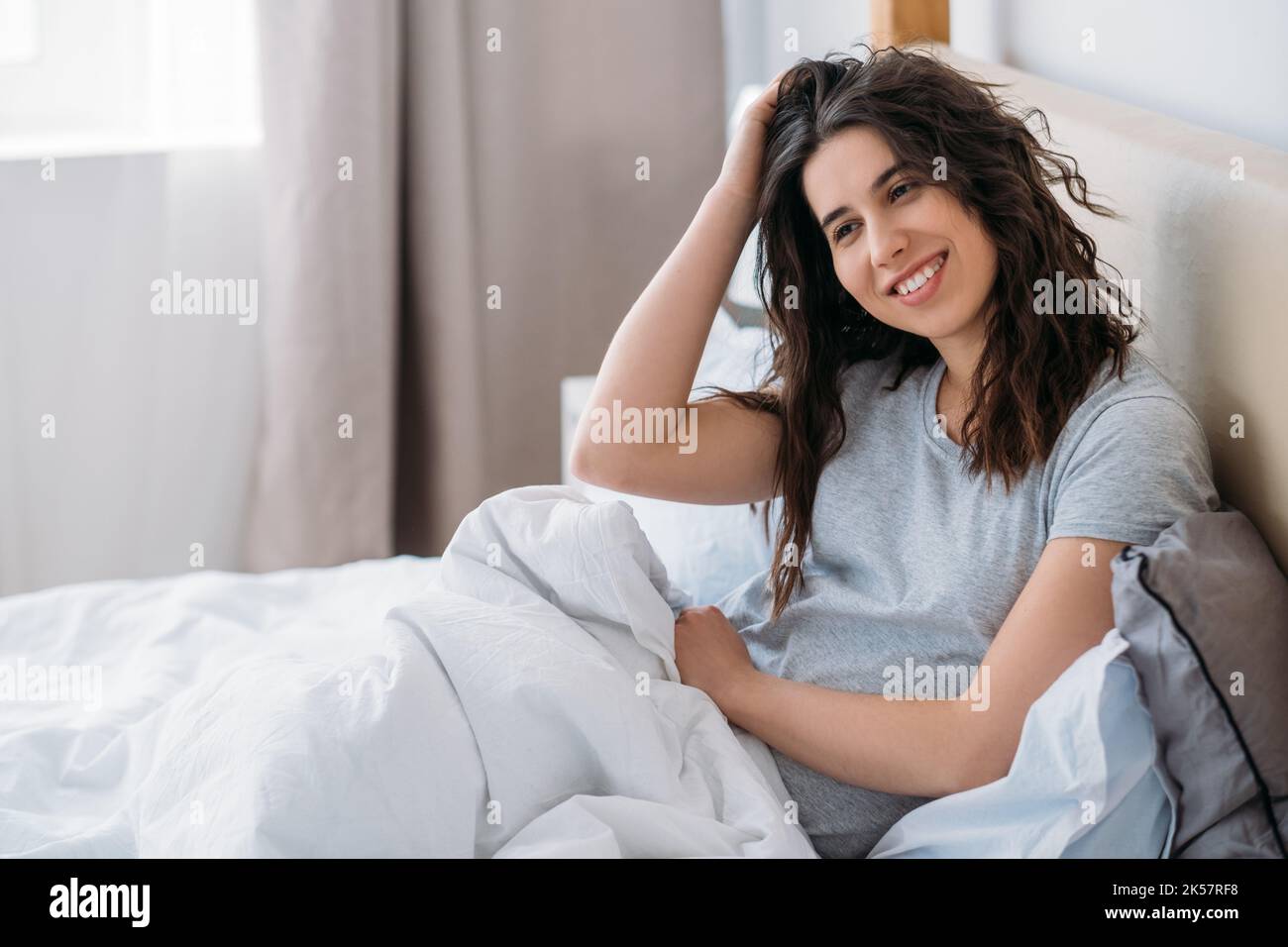 Lazy morning. Weekend relaxation. Home leisure. Happy cheerful brunette woman enjoying rest sitting awake in cozy bed smiling in light bedroom. Stock Photo