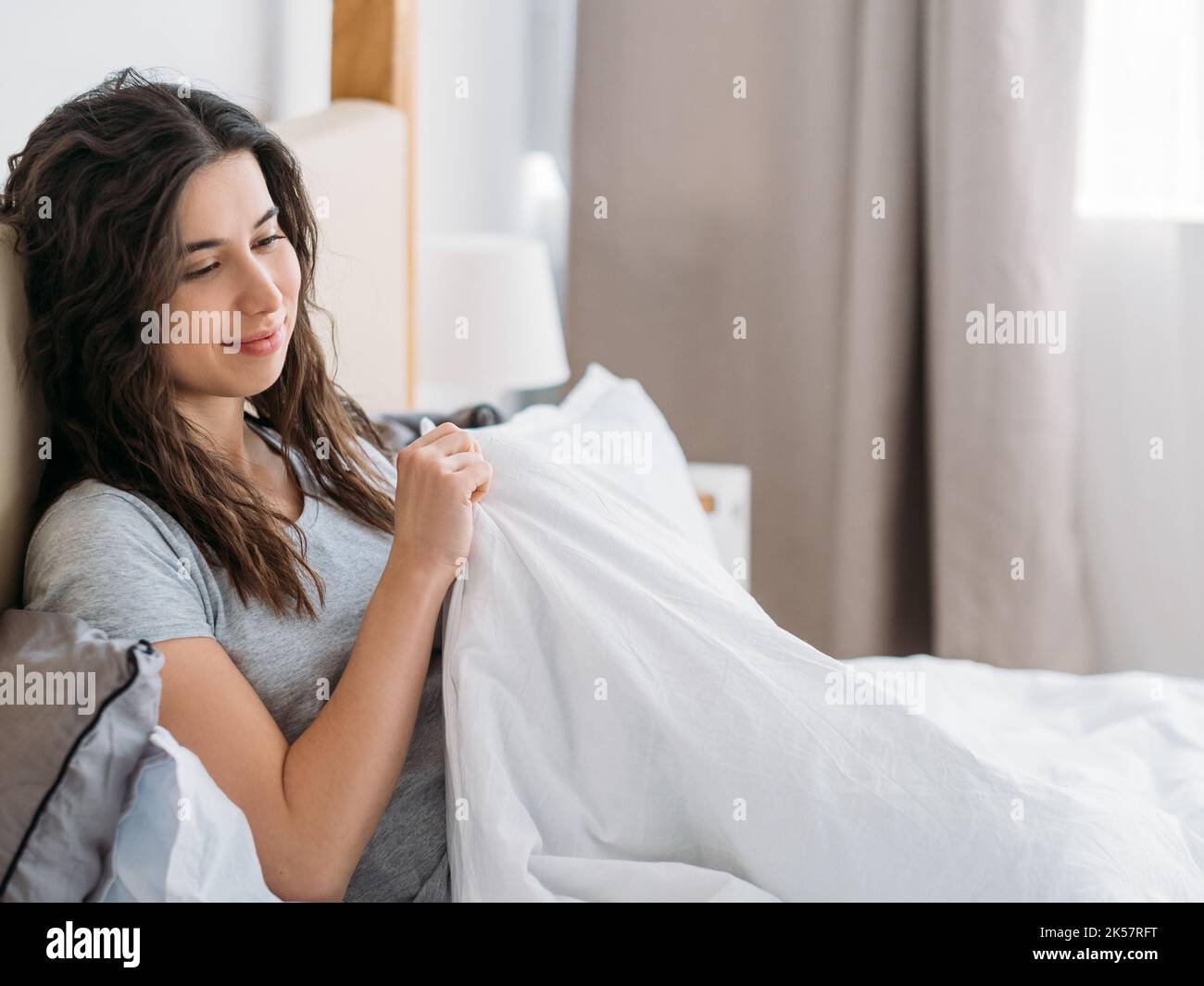 Romantic morning. Home rest. Pleasure inspiration. Relaxed pensive daydreaming brunette woman smiling in cozy bed with blanket in light bedroom. Stock Photo