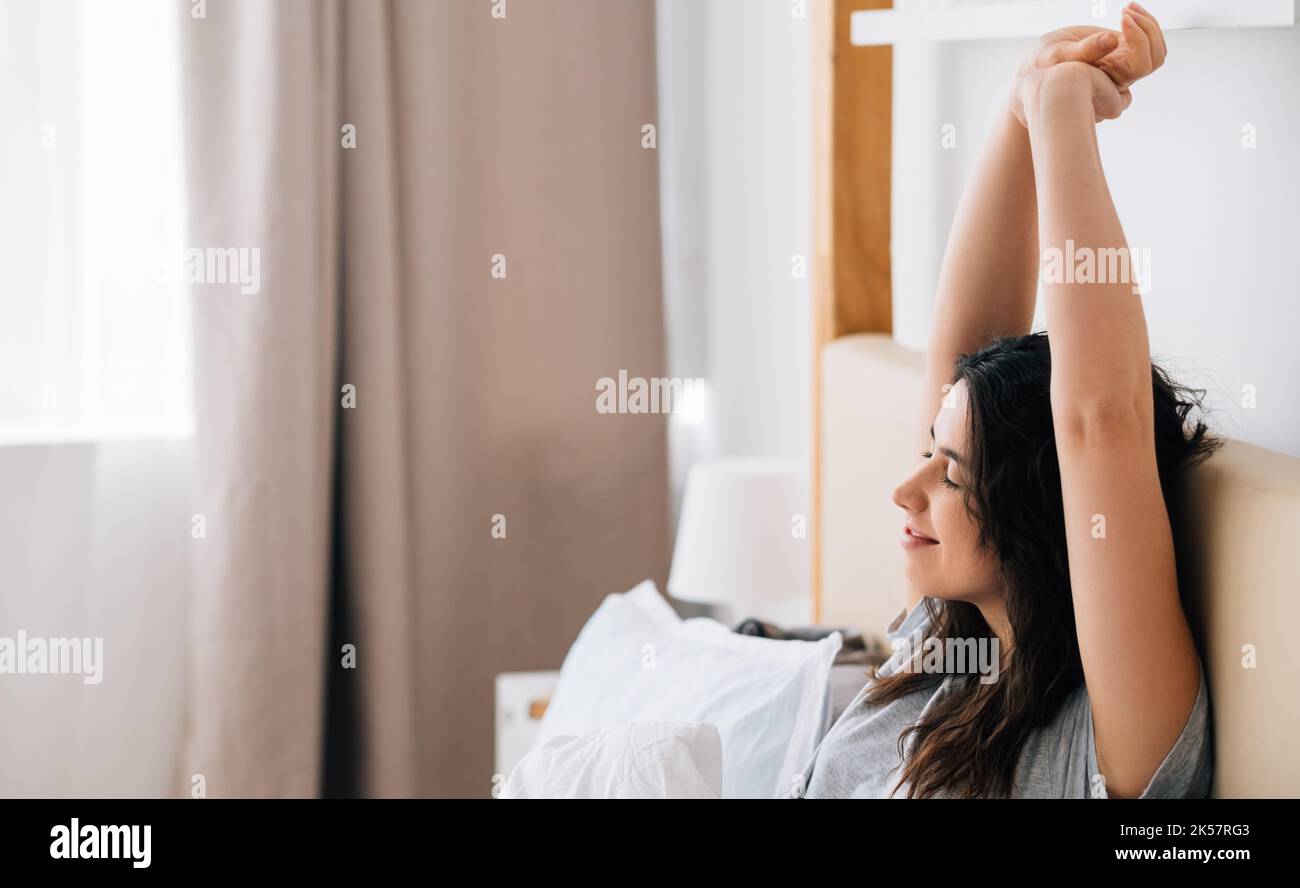 Good morning. Weekend leisure. New day beginning. Health wellbeing. Relaxed happy brunette woman stretching smiling in bed in light home bedroom. Stock Photo