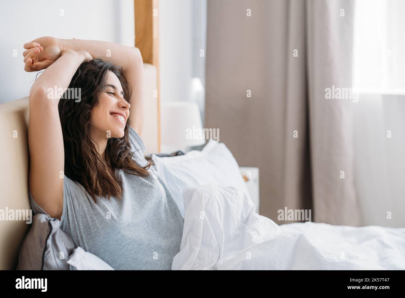 Happy morning. Home relaxation. Positive lifestyle. Satisfied cheerful brunette woman enjoying rest alone in cozy bed smiling in light bedroom. Stock Photo