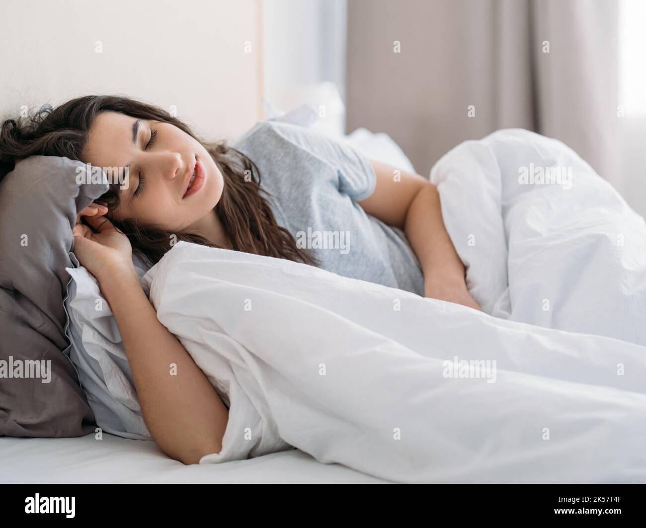 Morning nap. Lazy weekend. Home relaxation. Peaceful happy brunette woman sleeping enjoying rest lying in cozy bed with white blanket in light bedroom Stock Photo